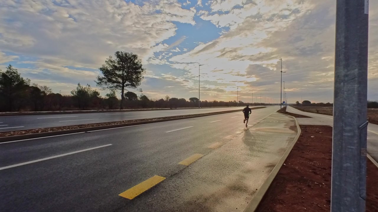 Wide shot of an athlete jogging away on a wet asphalt road toward the horizon. Dramatic morning sky and reflections. Concept of endurance, freedom, and personal journey.