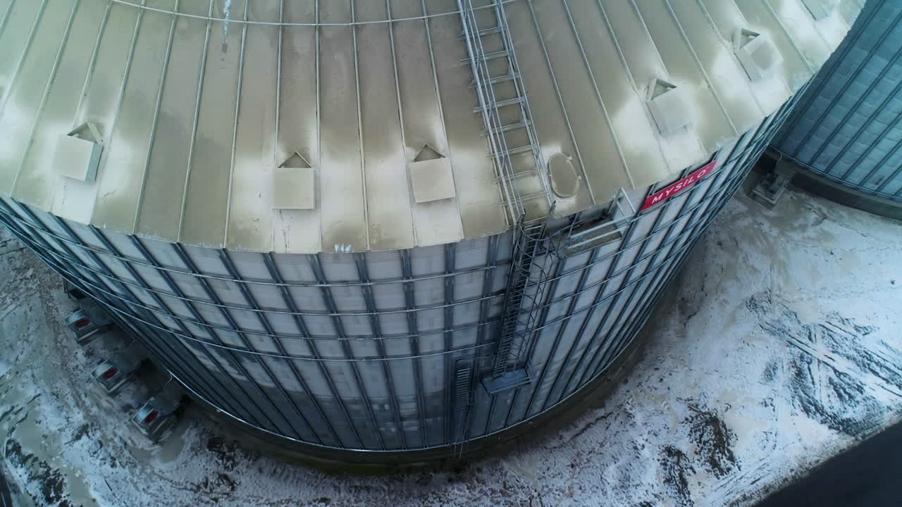 Flying around the enormous metal tank at agricultural elevator. Aerial footage over the dusty silo. Snowy ground backdrop.