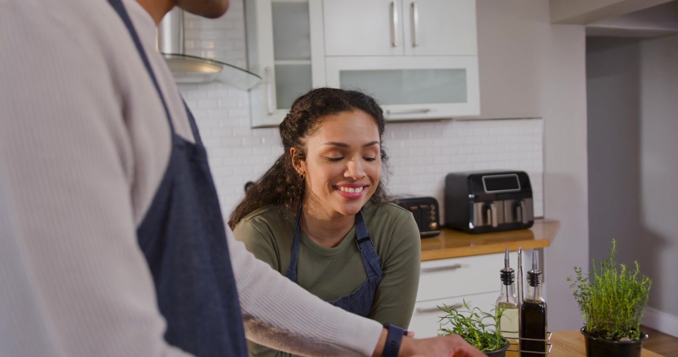 Coppia afroamericana in grembiule che osserva un uomo prendere un tagliere in cucina, mentre affetta l'impasto e cucina.