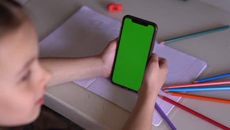 una niña está sentada en una mesa con el teléfono en las manos, mira el croma