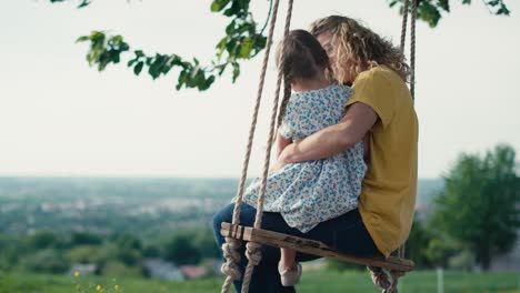 rear view of mom with little girl swinging and talking together in summer day.