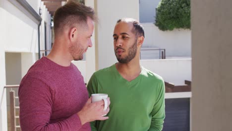 multi ethnic gay male couple drinking coffee on balcony in sun