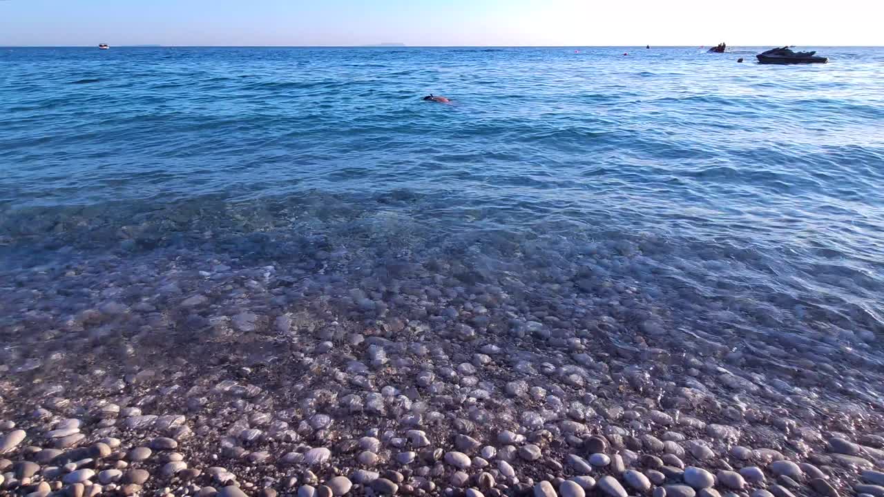 바다 물이 아름다운 해변에  ⁇  자갈을  ⁇ 고, 소년 스노클링과 모터 보트가 지중해의 조용한 물에서 떠다니고 있습니다.