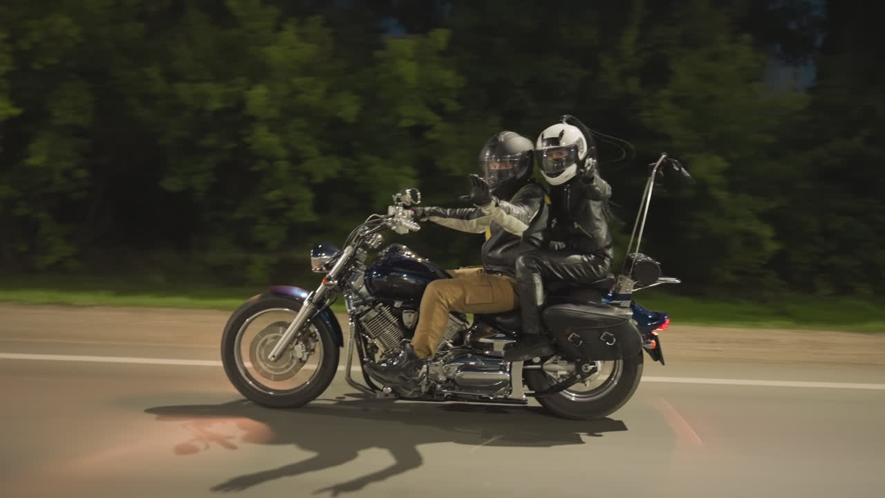 Side view of siblings riding motorcycle at night, both wearing helmets and leather gear, moving along road as they greet someone with friendly gesture while cruising past trees under dim light