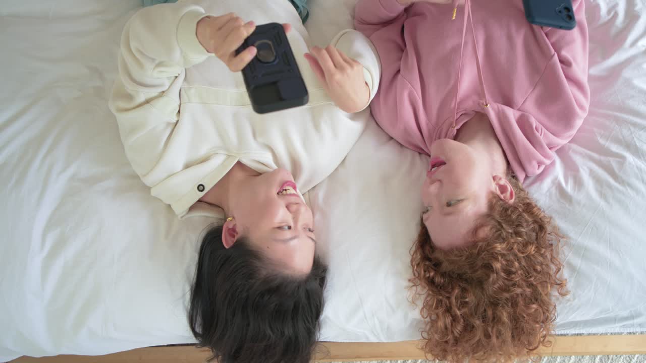 Den ene vennen løfter telefonen og viser innhold mens begge vennene ler og prater på sengen.
