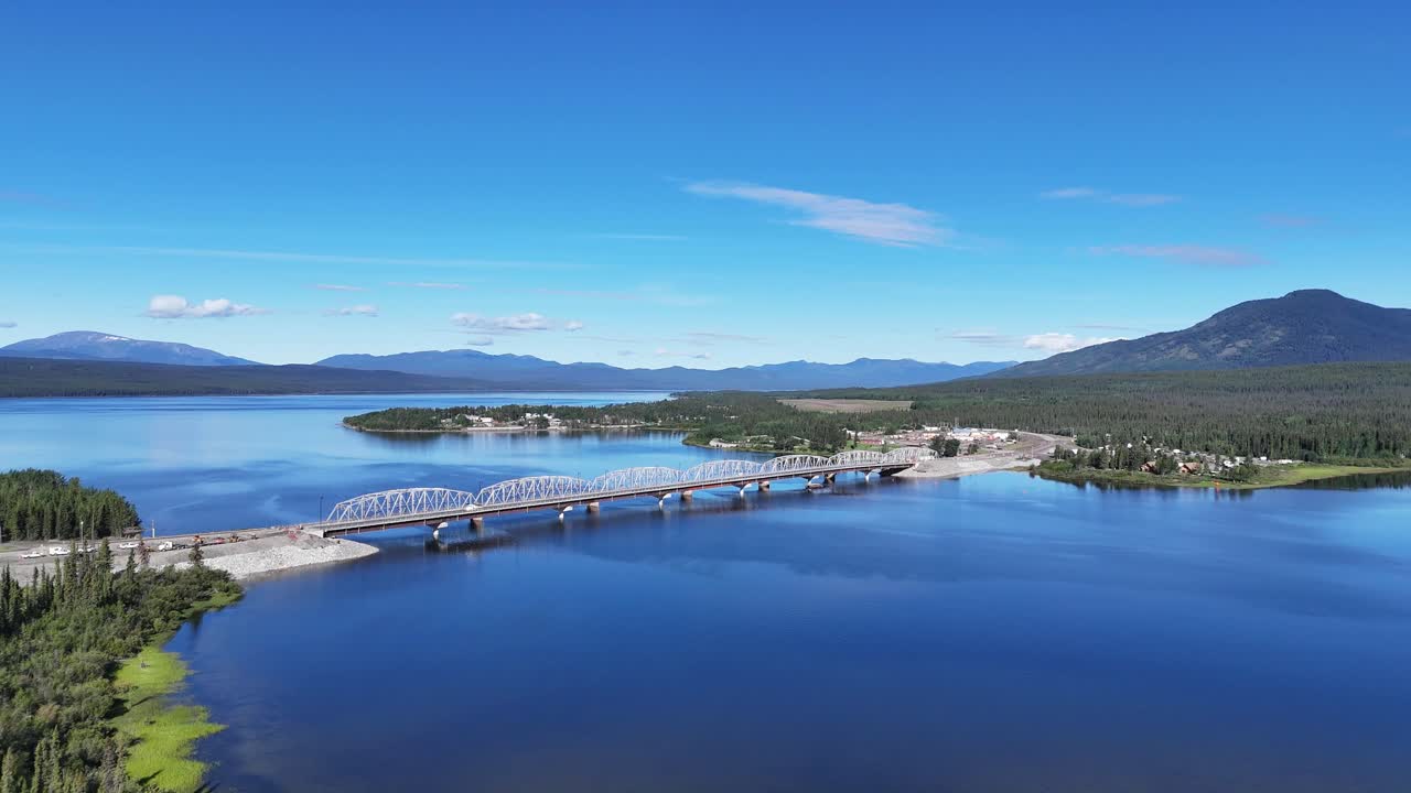 An aerial perspective captures a majestic bridge spanning a vast, tranquil blue lake, flanked by lush green forests and distant, hazy mountains under a clear, sunny sky in Canada