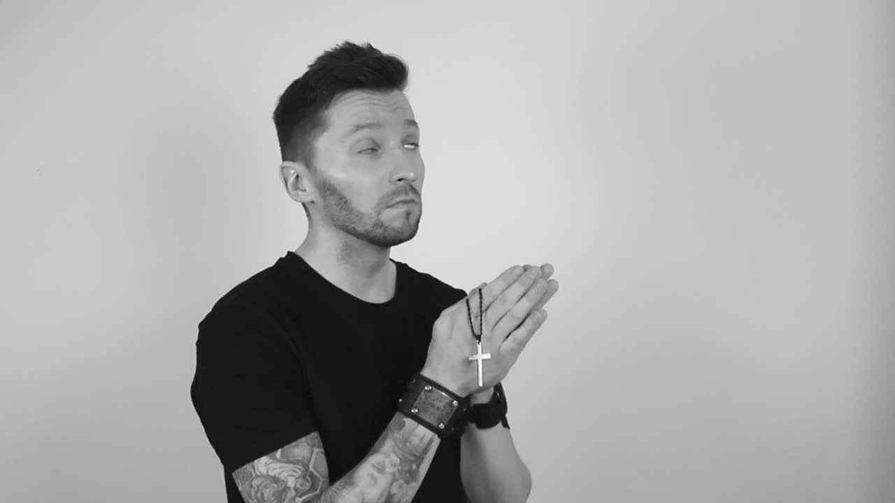 Grayscale footage of a tattooed man with short hair and a beard holding a small black holy cross necklace and prays with his hands together and closes his eyes with concentration. Religious man