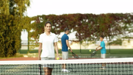 retrato de una hermosa mujer acercándose a la cámara con raqueta en la mano, apoyándose en la red y sonriendo a la cámara mientras pasa tiempo con su familia en una cancha de tenis