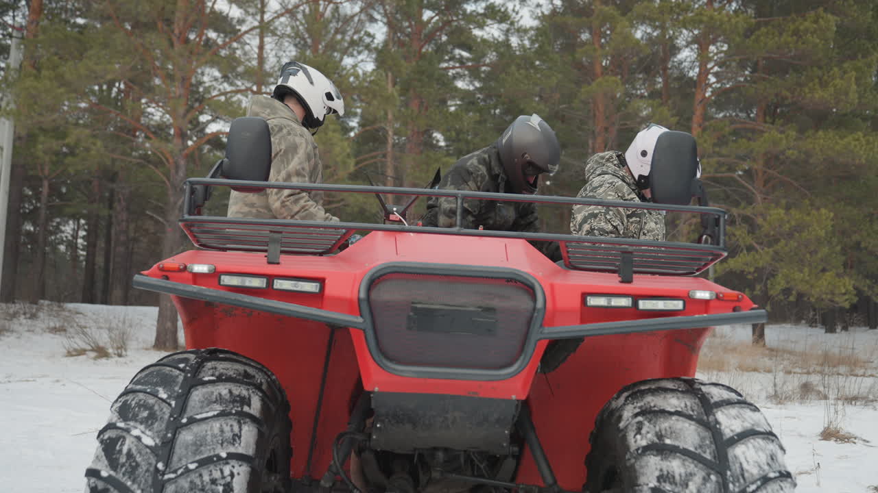 Equipaggio di ATV rosso che si prepara sulla neve, piloti con casco in mimetica che controllano i comandi, pneumatici da neve sovradimensionati, sfondo di pineta, istruttore che guida il passeggero sul sedile, atmosfera fredda invernale, lavoro di squadra