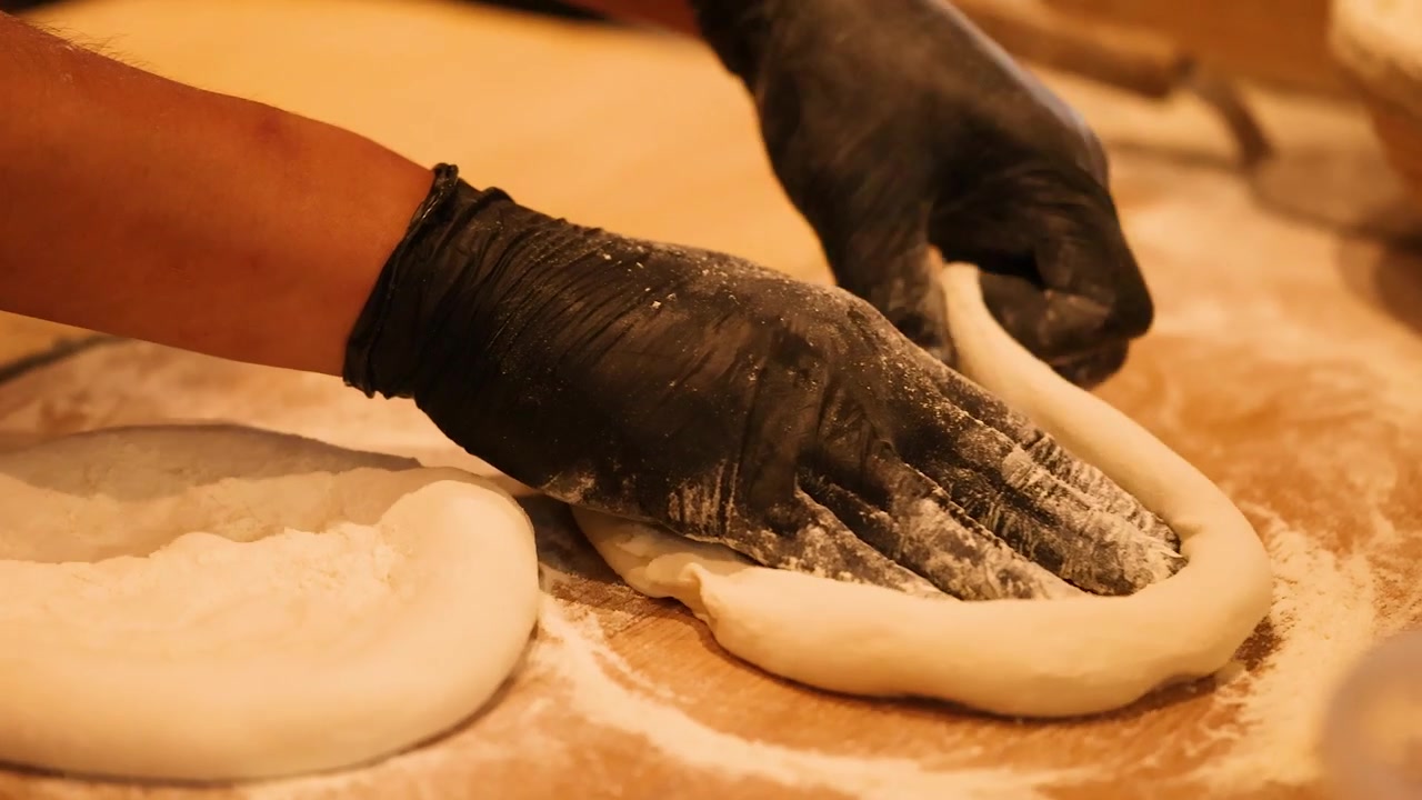 Primo piano di mani con guanti neri che stendono e impastano l'impasto della pizza su una superficie infarinata.
