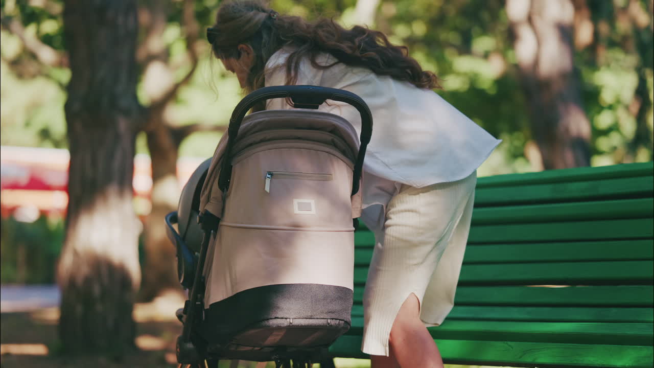 Young mother sitting on a park bench next to a stroller, enjoying a peaceful outdoor moment with her baby