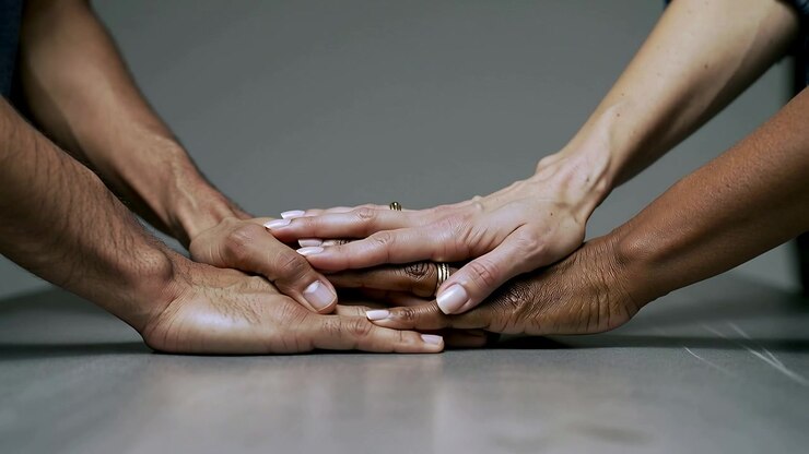 Reaching hands stacking on grey table and backdrop, showing unity with wedding ring and white nails