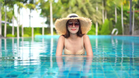 smiling-girl-chest-deep-in-a-beautiful-blue-water-pool-wearing-a-tan-sunhat-and-bathing-suit-with-vibrant-green-foliage-and-trees-in-the-background