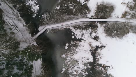 aerial view of person running over wooden path next to frozen lake with forest covered in snow