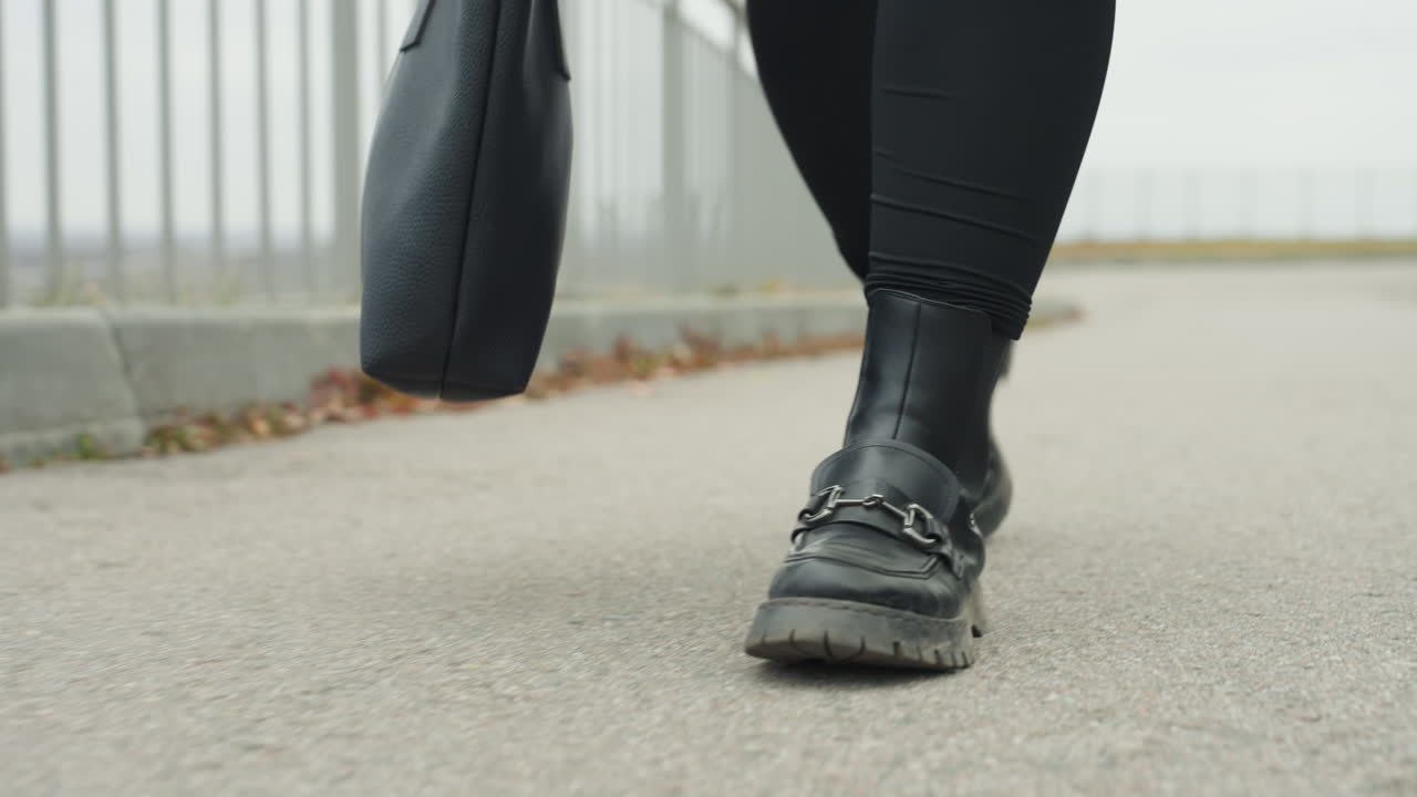 Primo piano di stivali che camminano con sicurezza sul marciapiede, stivali di pelle neri e borsa tote in un vivace tragitto urbano lungo la ringhiera di un ponte, passi ritmici ripresi da angolazione bassa, foglie autunnali sul bordo, cinematografico