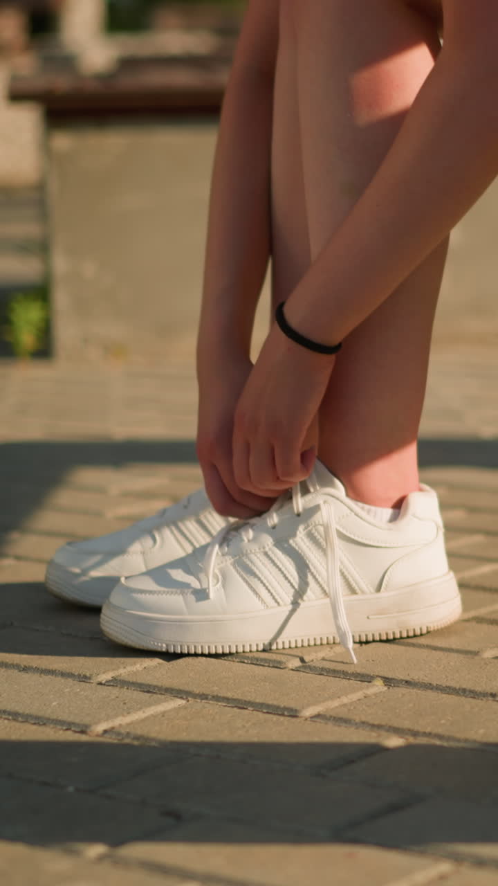 Lower view of individual loosening sneaker shoelace while seated on bench, wearing white sneakers and black bangle on right hand, sunlight casts shadow on ground