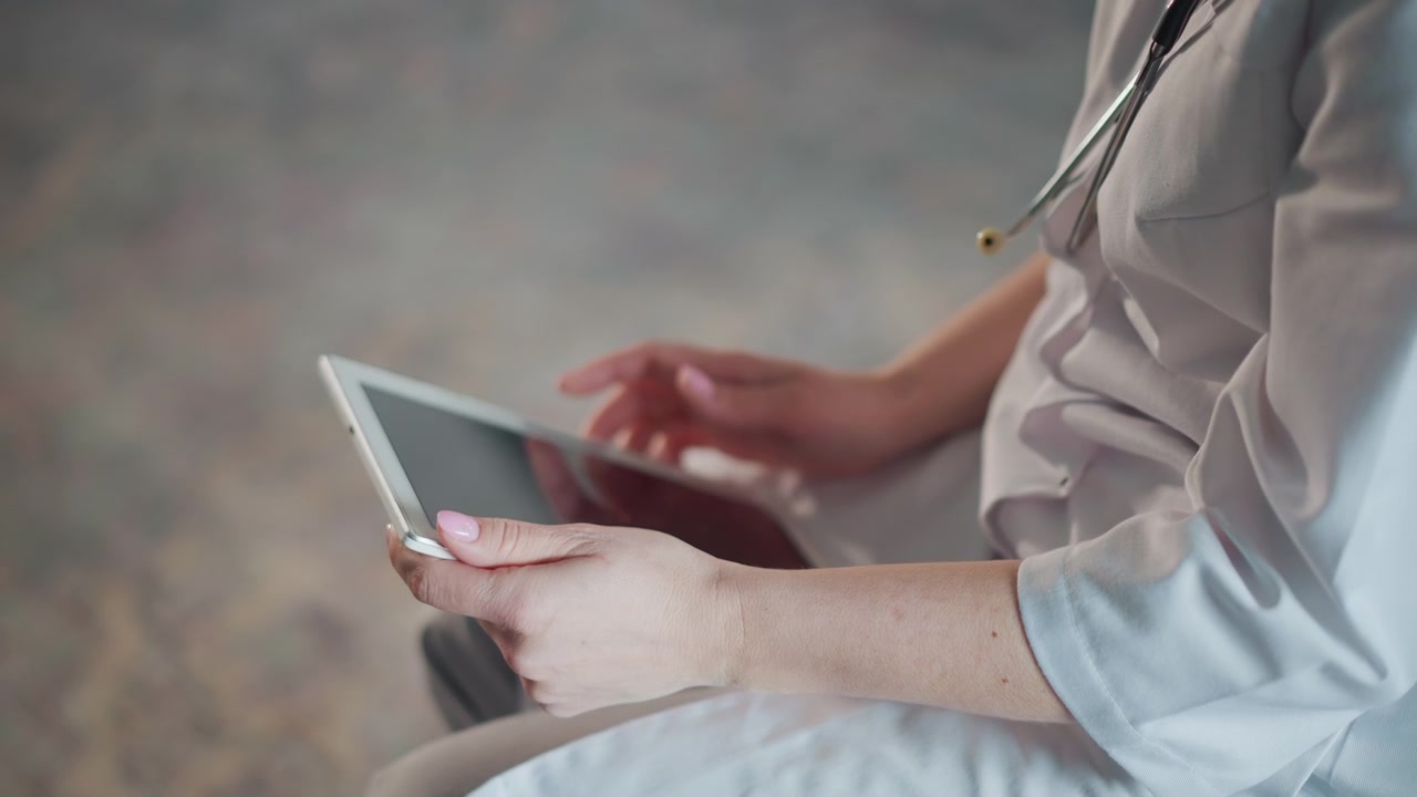 En tankefuld kaukasisk læge med tablet i et stille rum, i klinisk eftertanke. Stetoskopet er synligt, masken er sænket, og udtrykket er fokuseret, mens han/hun trykker på skærmen og studerer journaler på skødet i et afdæmpet interiør