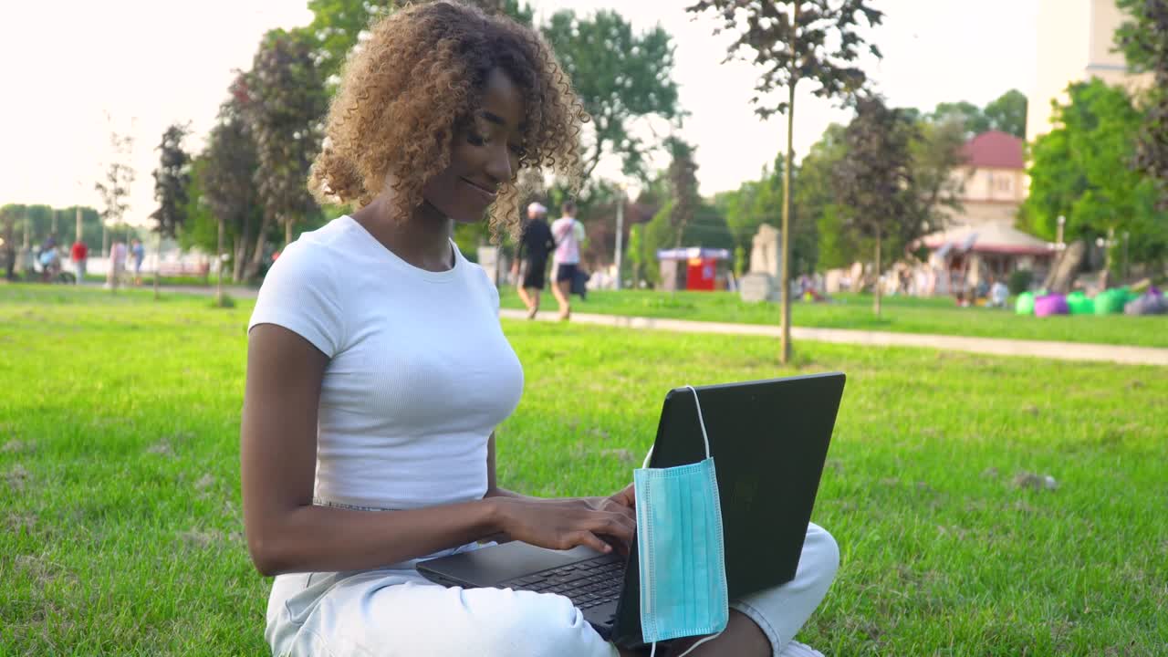 Young african american woman using laptop in park during covid 19 pandemic. Medical disposable mask hanging on display