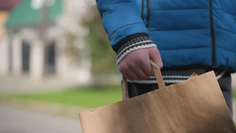 primer plano de la mano de un niño que sostiene una bolsa de papel mientras camina en una zona residencial las sombras se reflejan en él, con árboles de otoño vibrantes y un fondo borroso