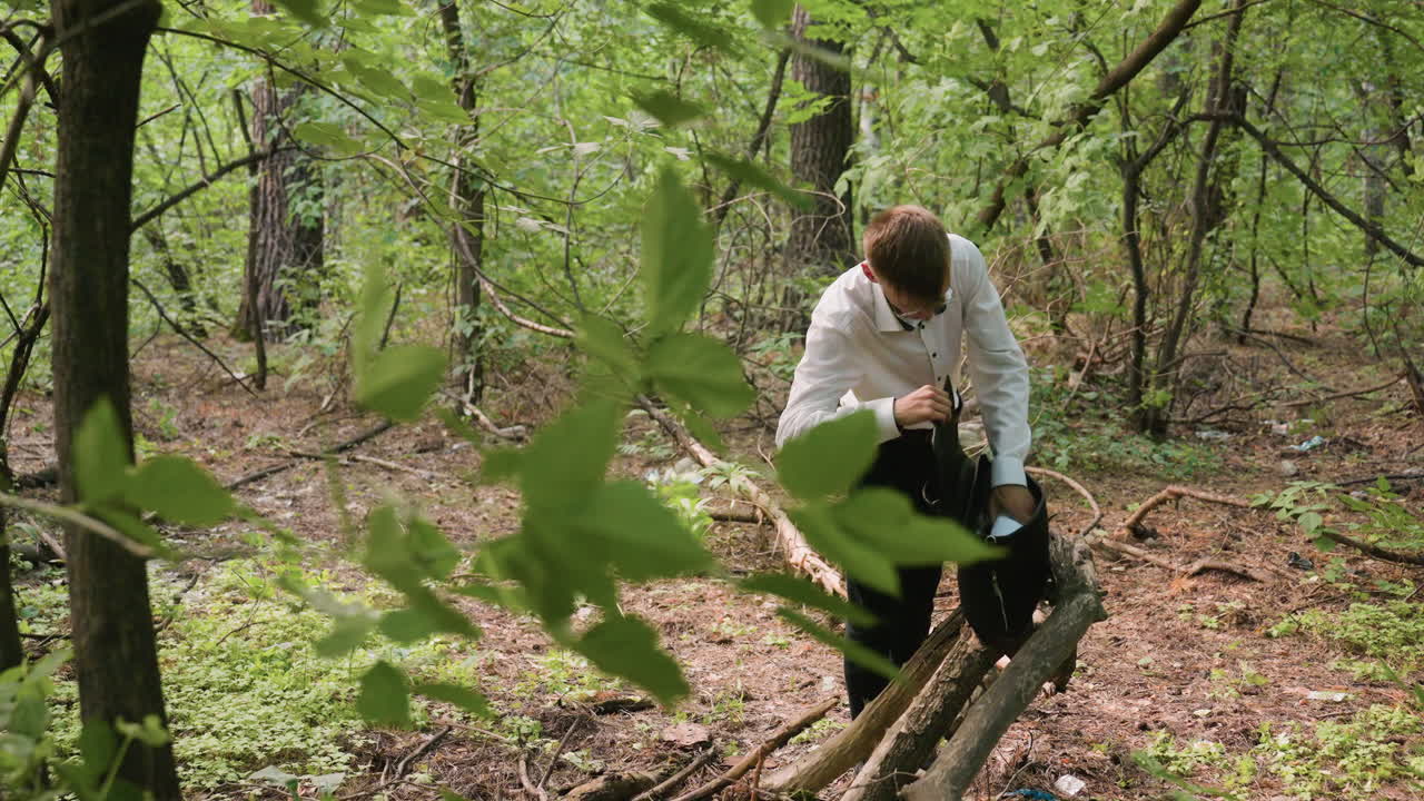 Scientific researcher in white shirt carefully returning white coat into black leather bag resting on tree branch in forest, surrounded by greenery, trunks, branches