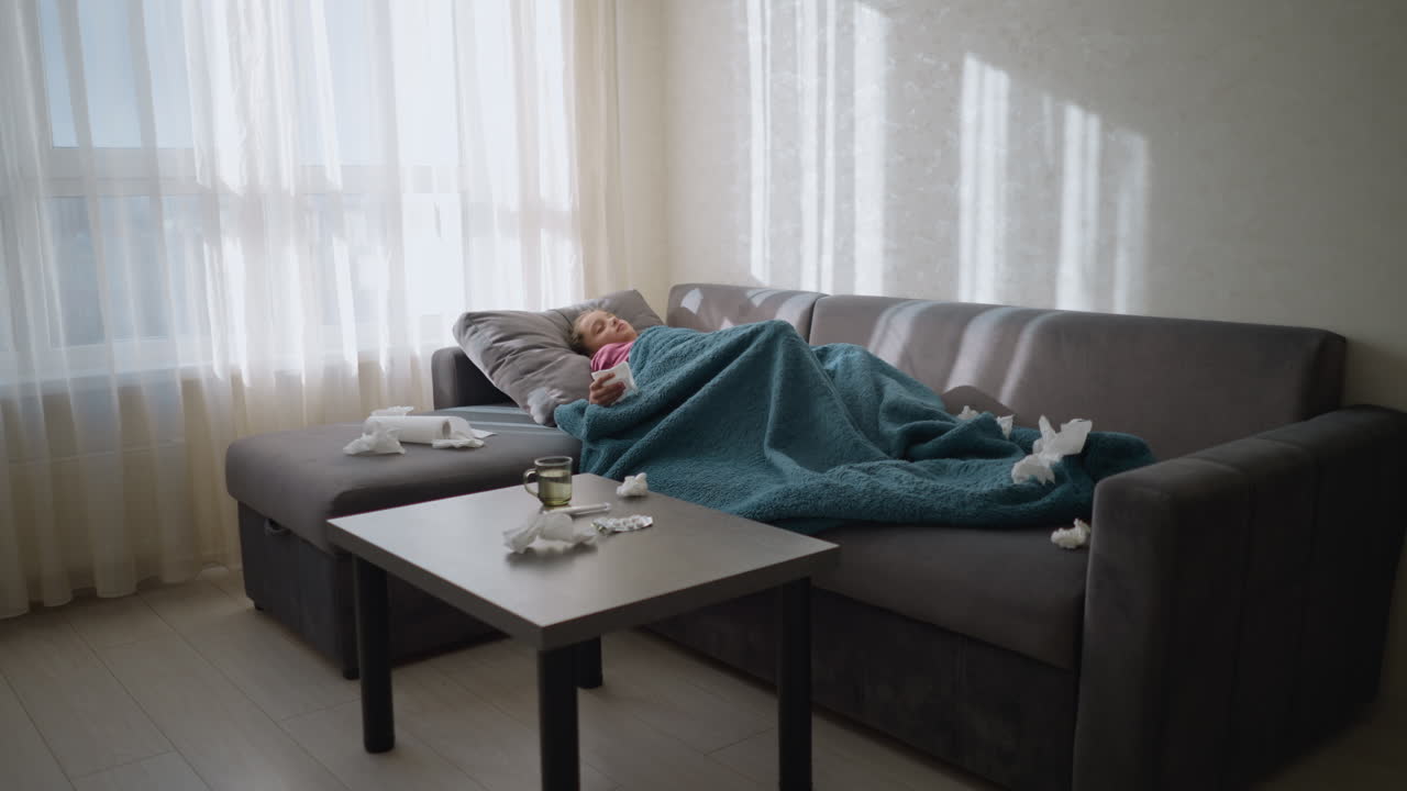Sick kid lying on couch covered with blanket adjusting herself, surrounded by tissue paper scattered around, flu symptoms visible with tissues, medicine, and drink on table in living room