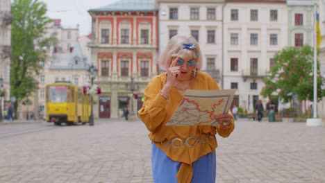 Senior-stylish-tourist-granny-woman-walking-along-street,-looking-for-way-using-paper-map-in-city