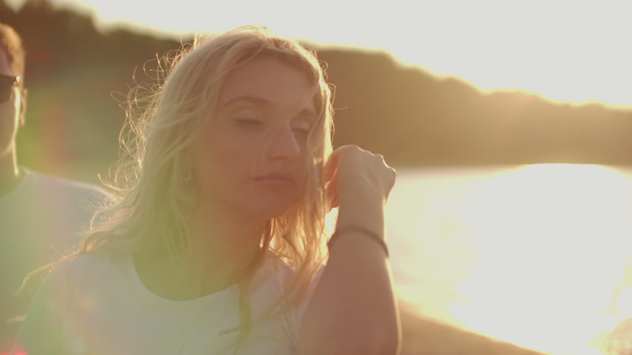A blonde woman student with bright makeup and long earrings enjoys dancing and party on the beach with beer. She touch her long hair and move her young body.