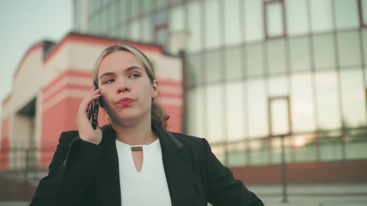 Frustrert hvit dame i profesjonelt antrekk uttrykker utmattelse mens hun er i en telefonsamtale i et urbant miljø, hånden hevet i stress, folk uskarpe i det fjerne bakgrunnen