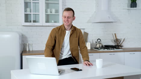 young man works and drinks tea in the kitchen. happy student studying at home during quarantine. guy using a laptop for job in comfortable apartment.