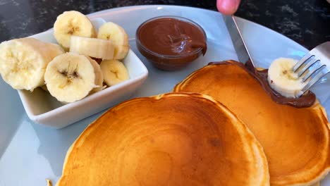 a person cutting golden pancakes with bananas and chocolate sauce on the side, tasty breakfast, 4k shot