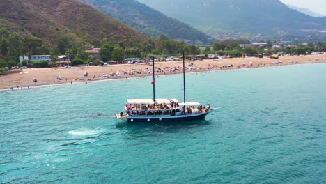 Luftdrohne-Umkreist-An-Einem-Sonnigen-Sommertag-Ein-Großes-Boot-Entlang-Der-Mittelmeerküste-Des-Strandes-Von-Adrasan-In-Der-Türkei-Voller-Touristen