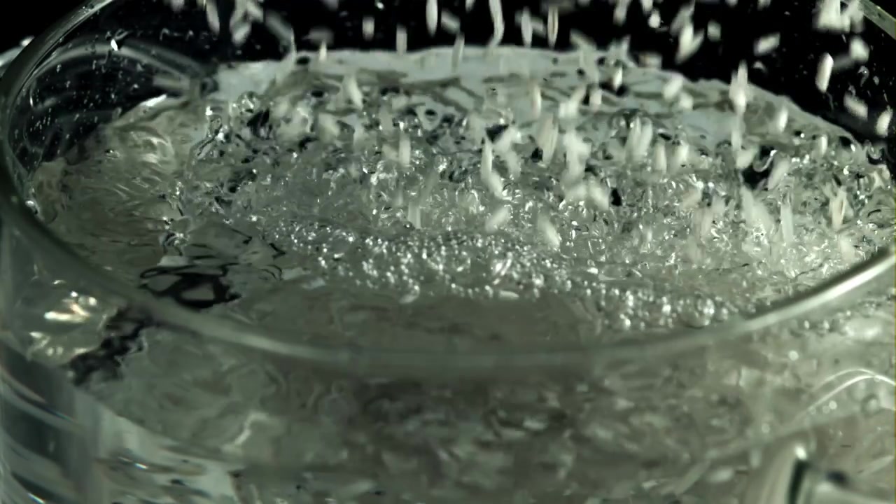 Versamento di grani di riso in acqua bollente in pentola di vetro al rallentatore