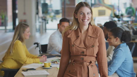 Portrait-Of-Businesswoman-With-Colleagues-In-Background-Sitting-Around-Table-In-Open-Plan-Office