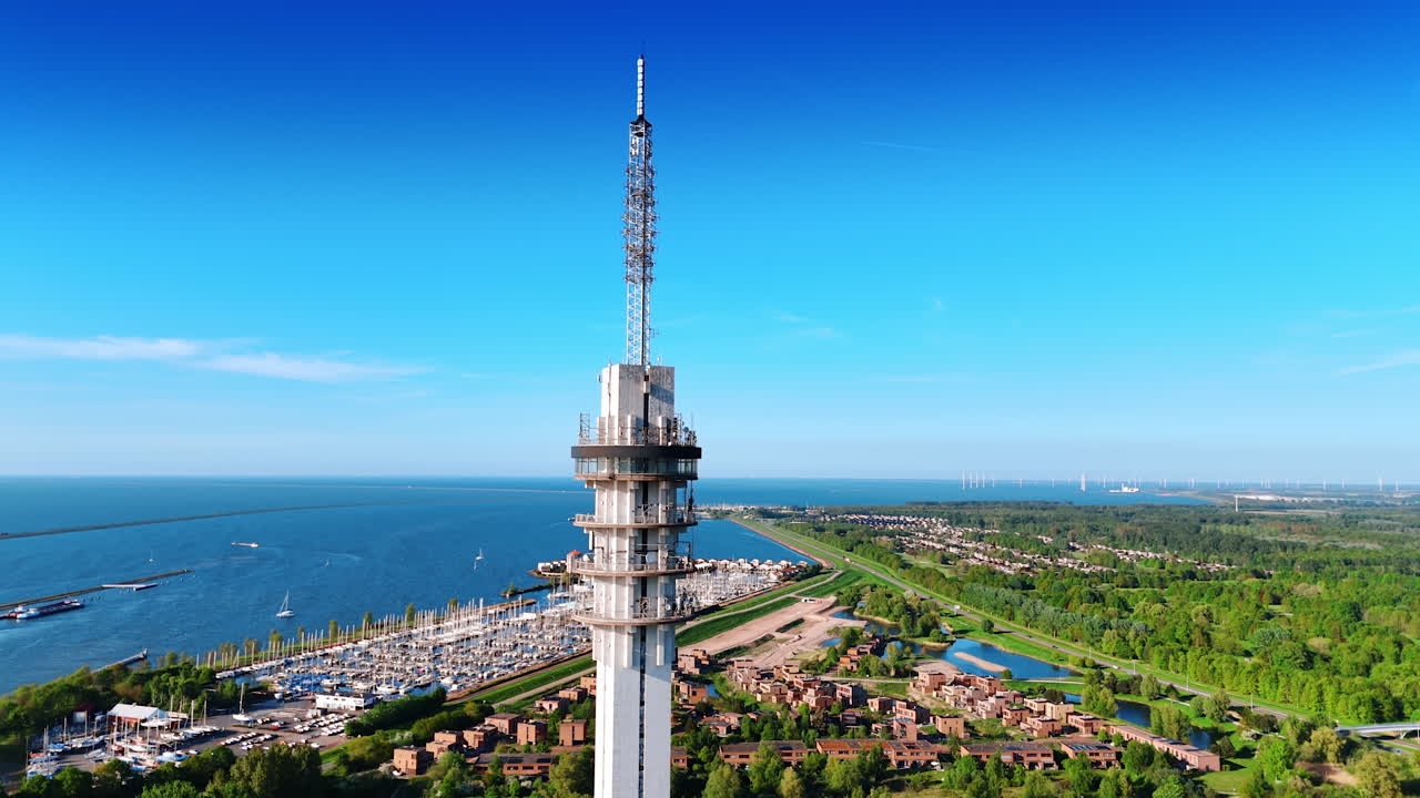 Top of the telecom tower at the backdrop of blue sky and azure waterscape of Houtrib Harbor, Lelystad, the Netherlands. Multiple yachts in the docks at backdrop. Aerial perspective.