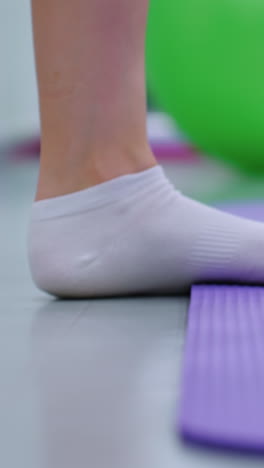 vista detallada de una entusiasta del fitness ajustando una banda de resistencia azul alrededor de sus tobillos en una alfombra púrpura, con una pelota de ejercicio verde suavemente borrosa en el fondo