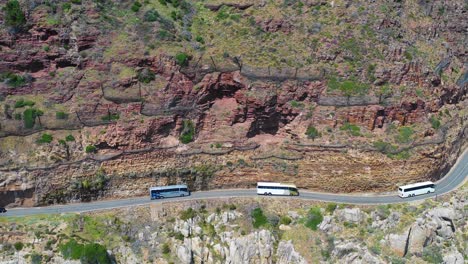 Una-Toma-Aérea-De-Un-Convoy-De-Autobuses-Que-Viajan-Por-Una-Peligrosa-Carretera-De-Montaña-Estrecha-A-Lo-Largo-Del-Océano-Chapmans-Peak-Road,-Cerca-De-Ciudad-Del-Cabo,-Sudáfrica-3