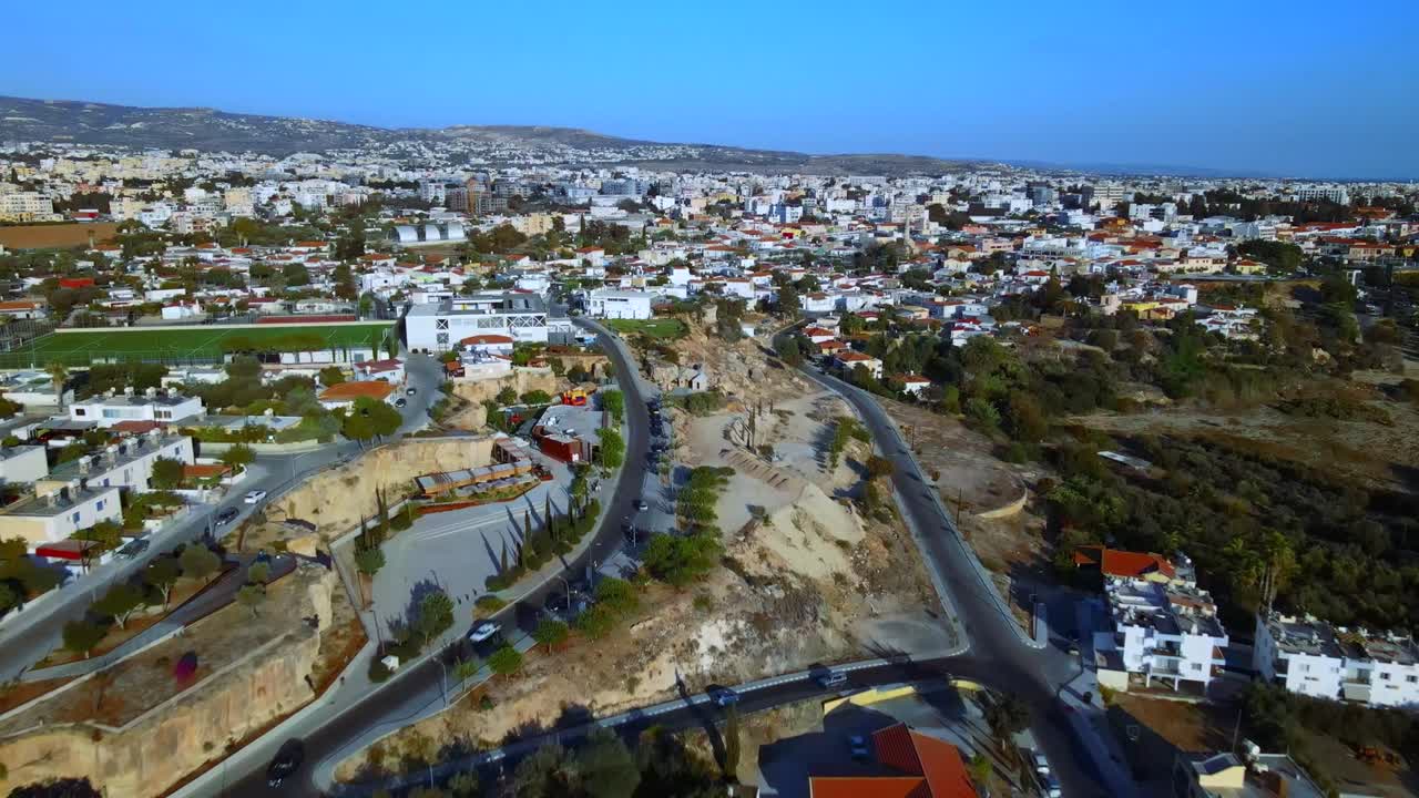 Drone view of hillside suburb with red-roof houses, winding roads, and wide city stretching to distant mountains under clear blue sky in peaceful daylight