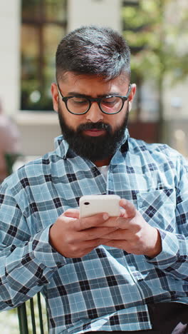 Indischer-Mann-Benutzt-Smartphone,-Schreibt-Textnachrichten,-Surft-Im-Internet-Und-Beendet-Seine-Arbeit-Mit-Blick-In-Die-Kamera