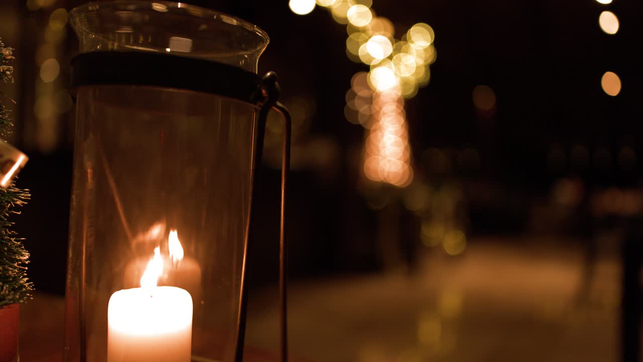Una candela bianca a colonna tremola all'interno di una lanterna cilindrica di vetro su uno sfondo di luci bokeh calde