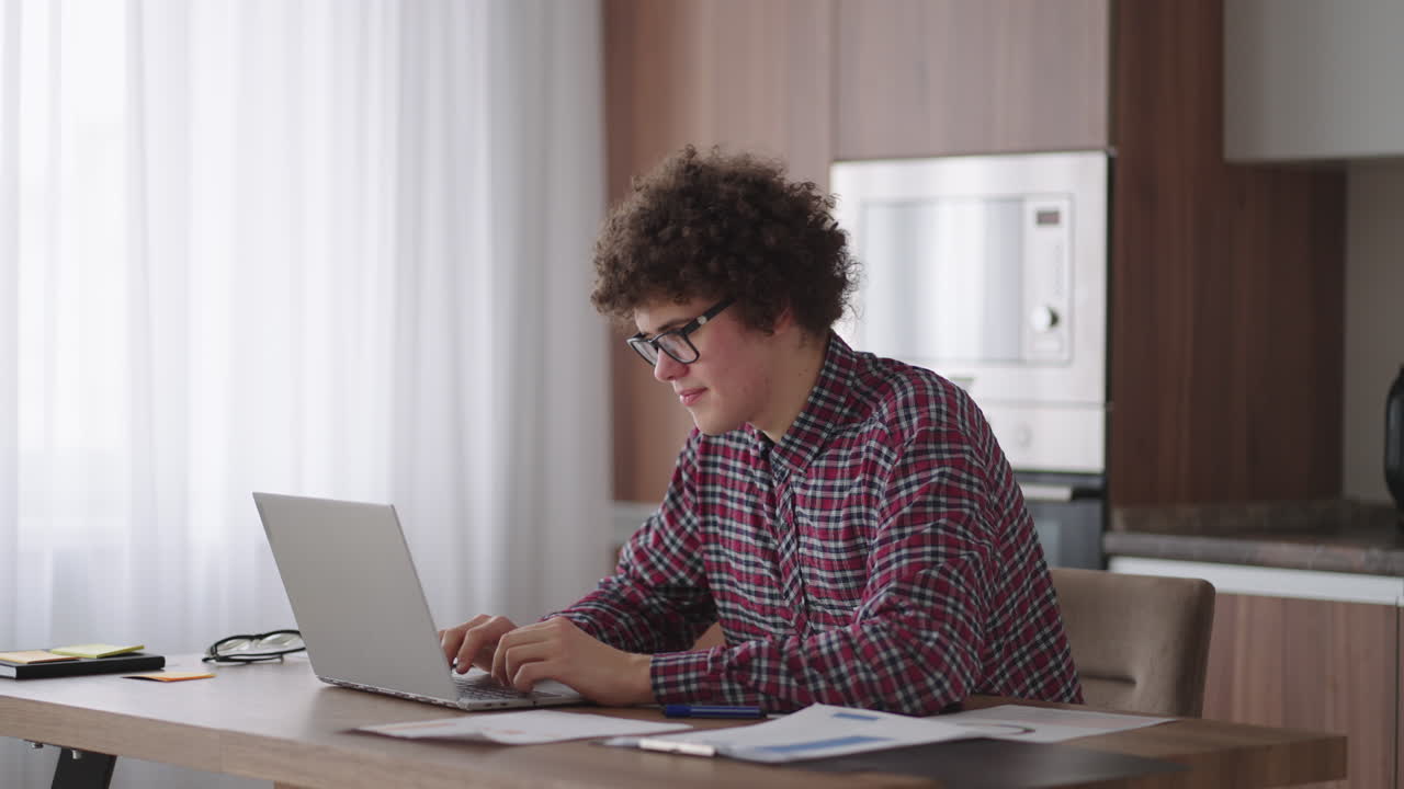 A Curly man with a serious look works at a laptop sitting in a modern kitchen. Young man freelancer student using laptop studying online working from home in internet