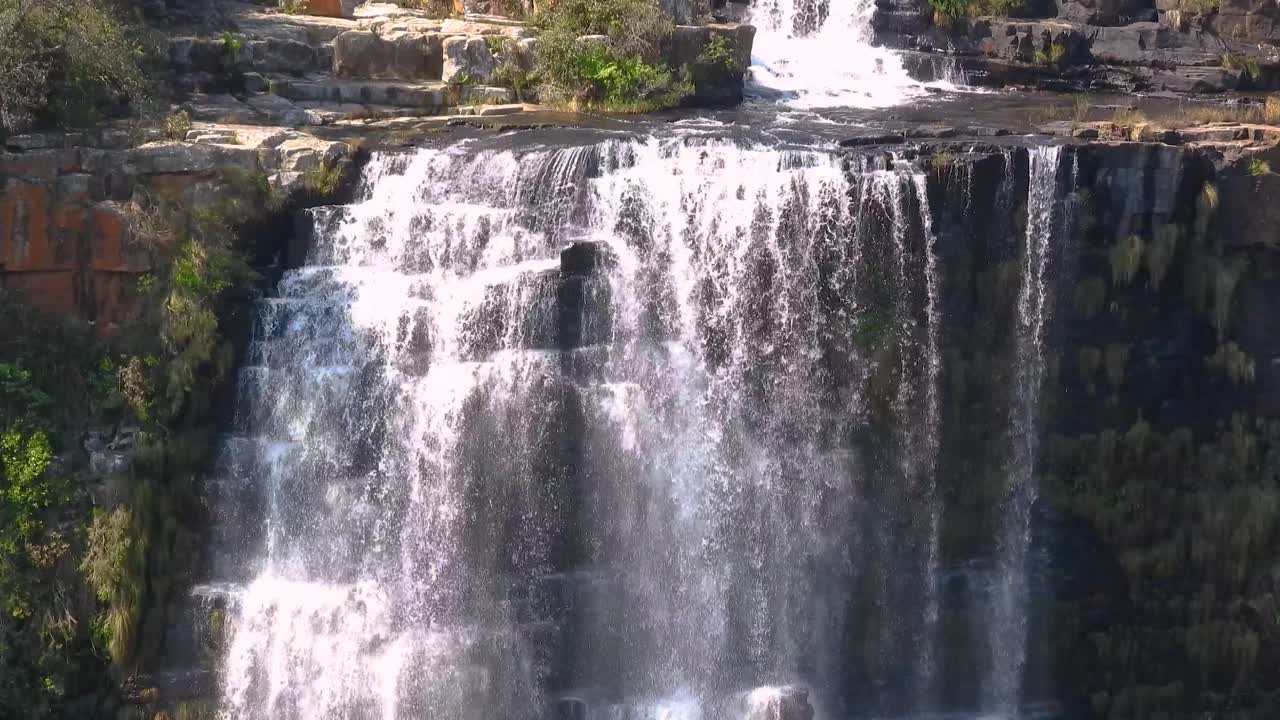 A stunning waterfall tumbles down rugged cliffs, creating a misty display as it crashes into the rocks below. Lush green vegetation surrounds the cascading water, highlighting the beauty of nature.