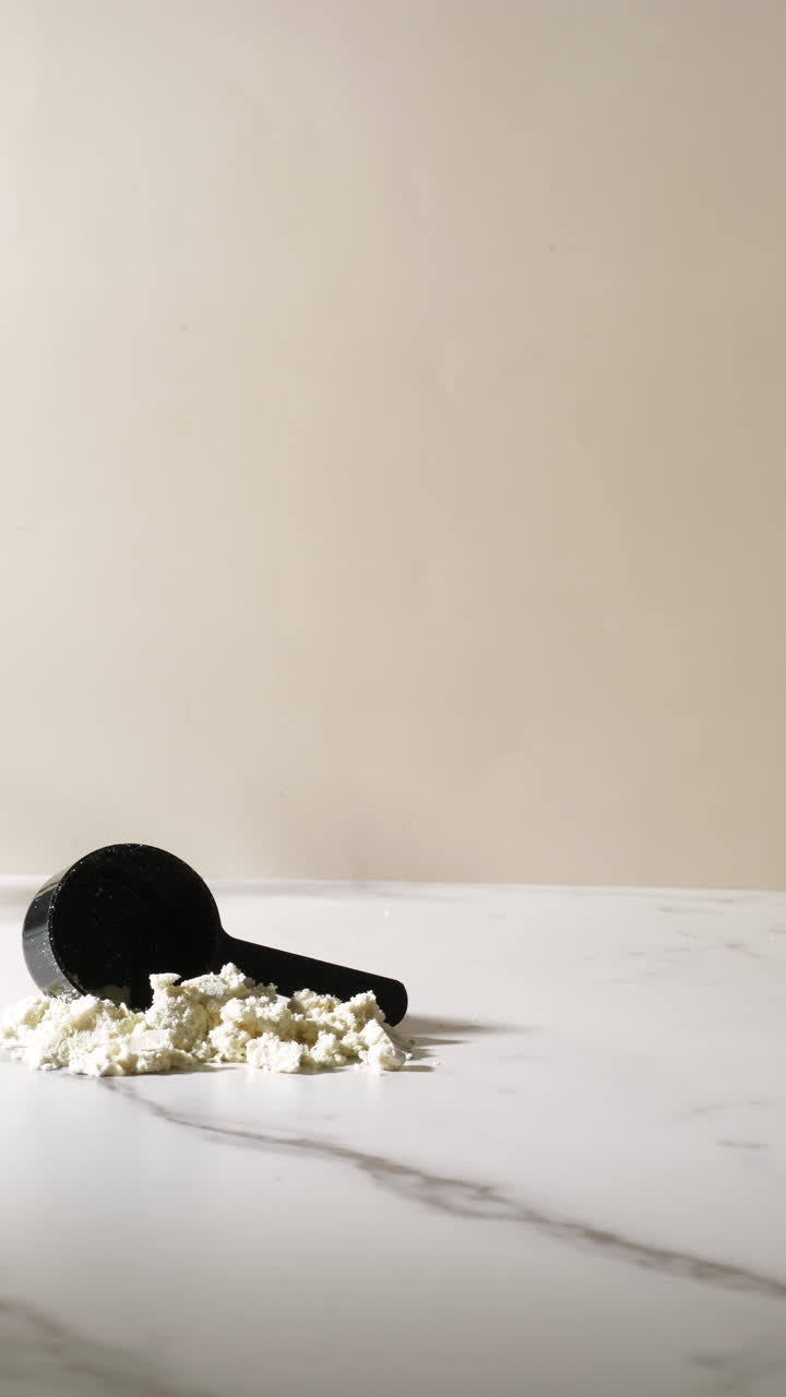 A hand lifts a wavy glass of creamy vanilla or banana protein shake from a marble surface, leaving behind a scoop and powder pile, captured in soft natural light with a clean, minimal beige backdrop.