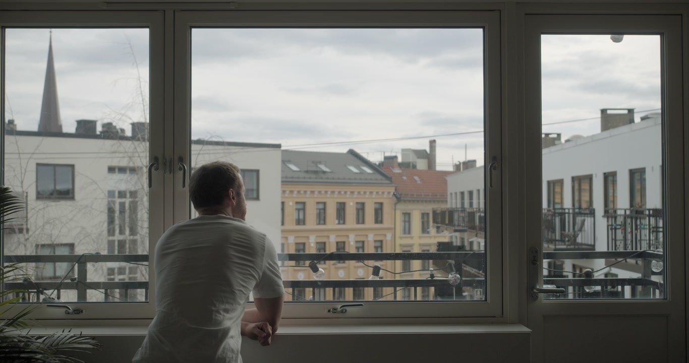 Tomada de câmera cinematográfica ampla e estática de costas de um jovem norueguês sozinho e isolado em quarentena de coronavírus, olhando pela janela de um apartamento escuro com paisagem urbana ao fundo, na parte central de Oslo, na Noruega.