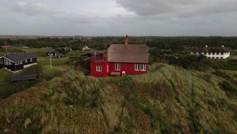 a drone orbits to the right around a red wooden house with a thatched roof at hvide sande beach, capturing the picturesque scene with stormy skies and sandy gras covered dunes in the background