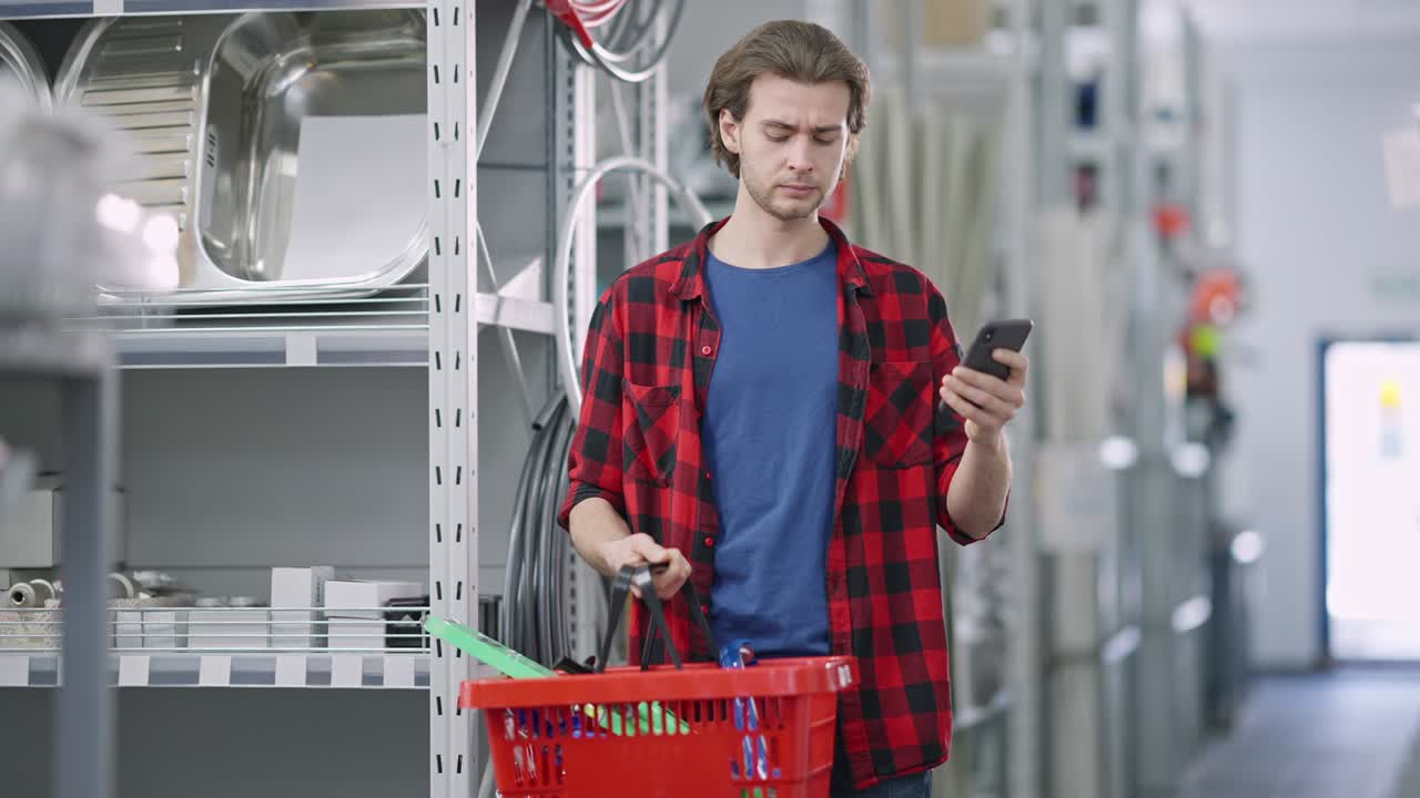 Concentrated young man checking list of purchases in smartphone with goods in shopping basket standing in hardware store. Portrait of confident male Caucasian buyer indoors. Industrial concept.