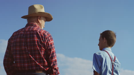 rear view of a grandfather in a hat and cute teen grandson talking outdoors with blue sky on the background