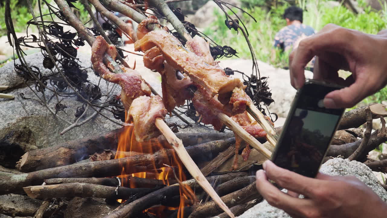 Cozinhando carne na fogueira na selva com outro cara tirando uma foto com seu iPhone