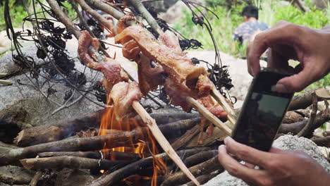 Kochen-Von-Fleisch-In-Flammen-Im-Dschungel-Mit-Einem-Anderen-Mann,-Der-Mit-Seinem-IPhone-Ein-Foto-Macht