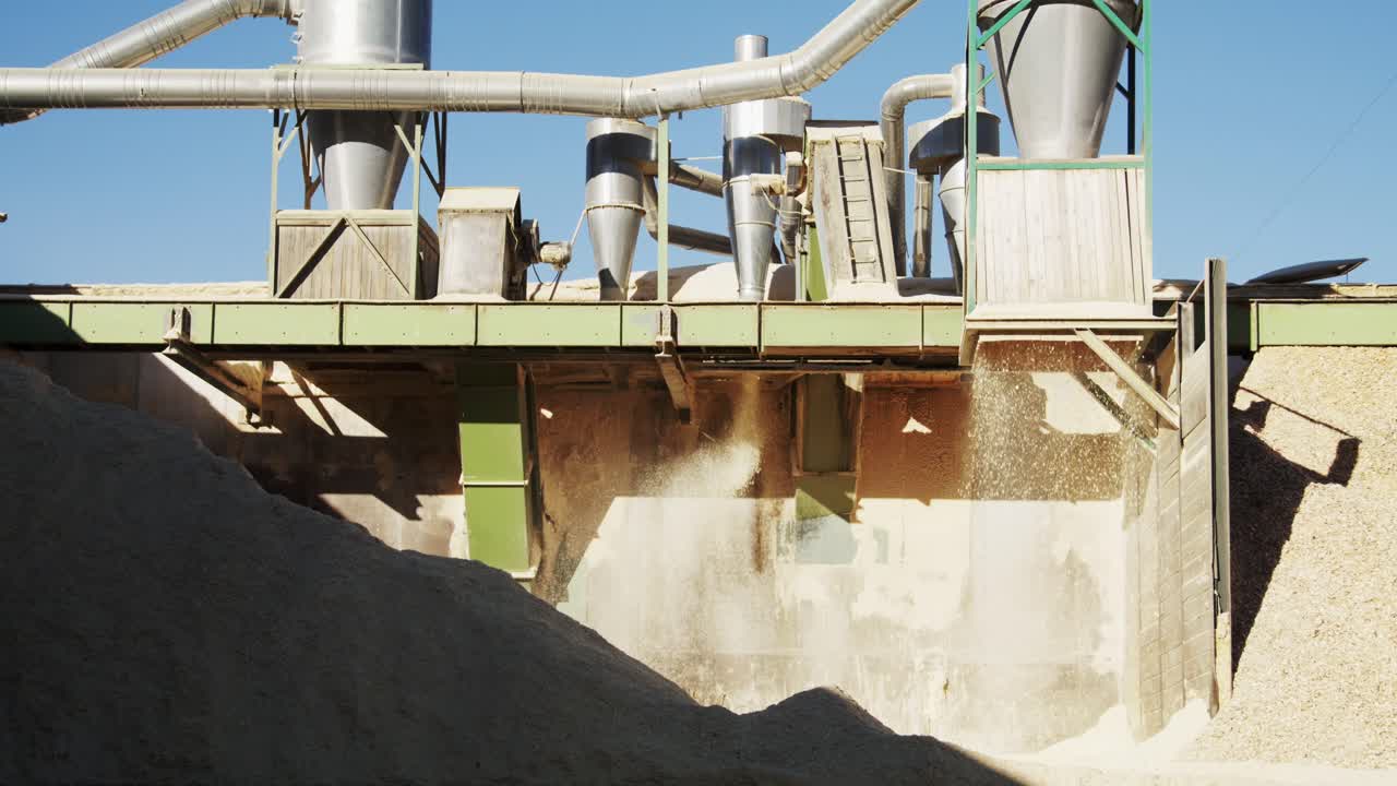 Industrielle maskiner på et savværk udleder en konstant strøm af savsmuld og træflis. Dette biprodukt fra træproduktionen opsamles i en stor bunke til genbrug eller biomasseenergi.