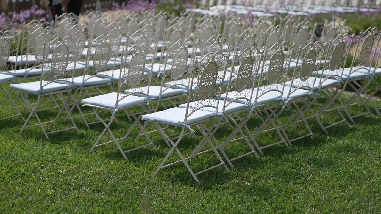Sedie in metallo bianche e ornate, vuote, disposte in file su prato verde per un matrimonio in giardino.